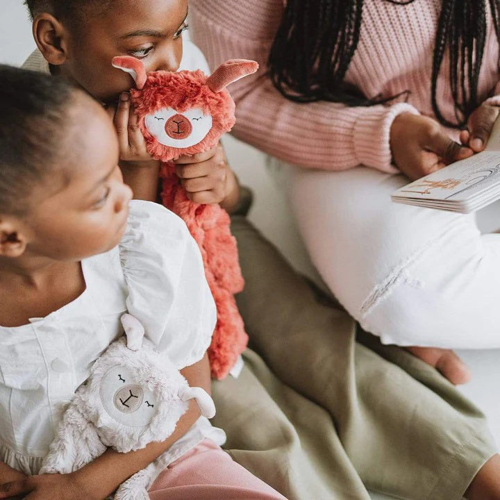 A cuddly SLUMBERKINS Alpaca Snuggler in rust is nestled in a white box adorned with the SLUMBERKINS logo. It includes a book titled "Alpaca," aimed at enhancing early emotional learning and managing anxiety.