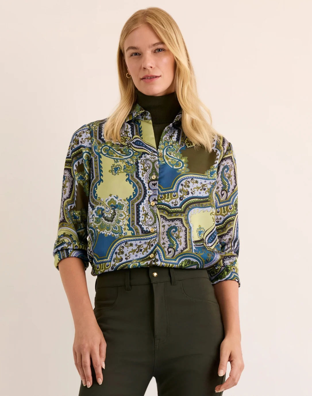 A woman with blonde hair wears the HINSON WU Margot Long Sleeve Paisley Shirt, a colorful button-up featuring green, blue, and yellow paisley patterns and crafted from luxurious Italian polyester for an elegant drape and silky feel. She pairs it with dark green pants and stands against a neutral background.