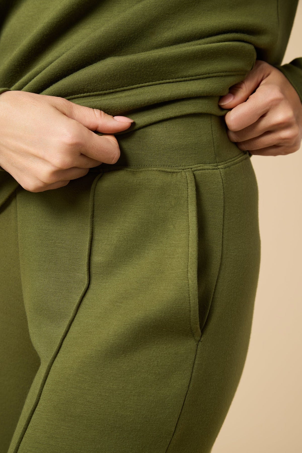 A woman wears a SOFTIES - DREAMTECH BOAT NECK WITH PINTUCK PANT SET in olive green, featuring a long-sleeve boat neck top and matching wide-leg pintuck pants.
