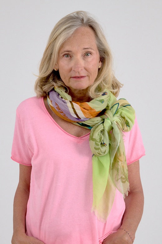 A blonde woman wearing a pink T-shirt, white shorts, and the STORIATIPIC - GERTRUD SCARF IN ANISE by STORIATIPIC stands against a plain white background, looking to her left.
