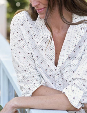 A woman wearing the FRANK & EILEEN Eileen Relaxed Button Up in Italian Cotton Palm Trees paired with blue jeans stands outside by a white building with large windows, smiling softly with one arm resting on her hip; the flattering button placement of the shirt by FRANK & EILEEN accentuates her silhouette.