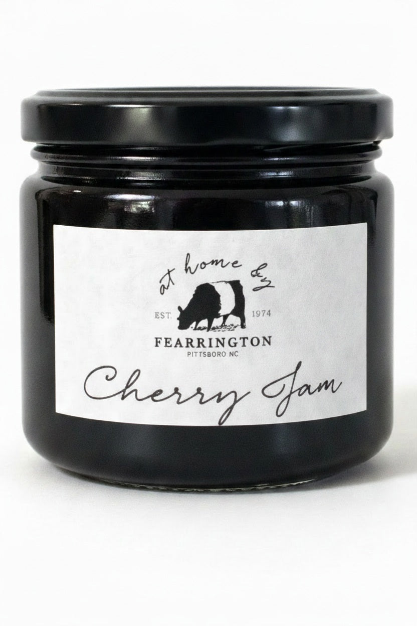 A glass jar with a black lid labeled “FEARRINGTON SIGNATURE COLLECTION - FEARRINGTON HOUSE CHERRY JAM” from Fearrington House, featuring a cow silhouette and the phrase “at home by Fearrington, Pittsboro, NC, EST. 1974,” on a white background. This versatile spread is crafted from ripe cherries to bring a deliciously sweet flavor to your pantry.