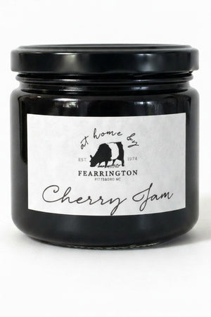 A glass jar with a black lid labeled “FEARRINGTON SIGNATURE COLLECTION - FEARRINGTON HOUSE CHERRY JAM” from Fearrington House, featuring a cow silhouette and the phrase “at home by Fearrington, Pittsboro, NC, EST. 1974,” on a white background. This versatile spread is crafted from ripe cherries to bring a deliciously sweet flavor to your pantry.
