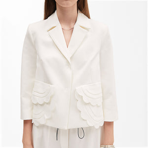 A person is wearing the MEIMEIJ - WHITE SHORT JACKET WITH SCALLOPED POCKETS, paired with a matching white top. The jacket, from the brand MEIMEIJ, features a notched collar and three-quarter sleeves. Complementing the outfit, they accessorize with a delicate necklace and a bracelet on their right wrist.