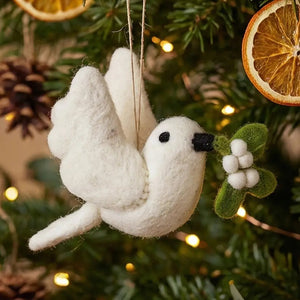 A MISTLETOE DOVE ORNAMENT by FELT SO GOOD hangs on a tree, the felt dove holding a sprig of mistletoe in its beak.