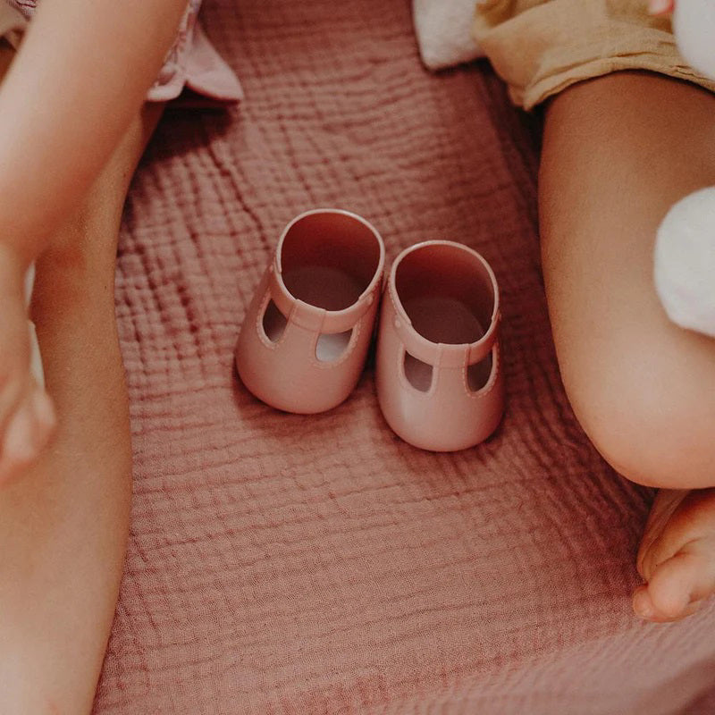 A close-up of a baby wearing OLLI ELLA USA DINKUM DOLL SHOES in Mallow Pink and white socks. The baby is seated on a soft, pale pink textured blanket, dressed in light brown clothing. Only the lower half of the baby's body is visible.