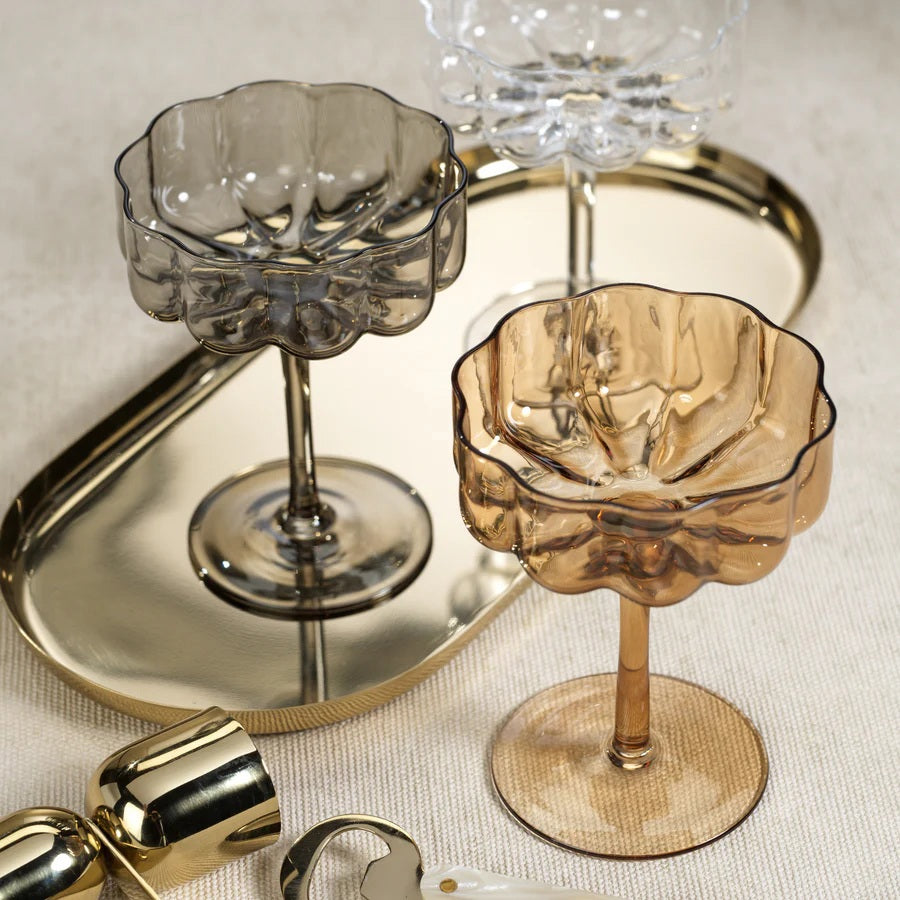 The ROCCO HANDBLOWN COUPE - AMBER by ZODAX features a brown-tinted, handblown amber glass with a scalloped, fluted bowl and a long stem, standing on a round base against a white background.