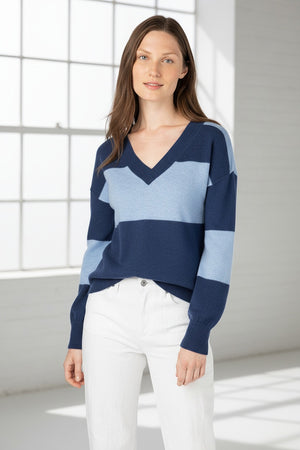 A woman with long brown hair models the KINROSS CASHMERE Gathered Sleeve Vee Neck pullover in pastel purple and peach, paired with white pants against a plain white background.