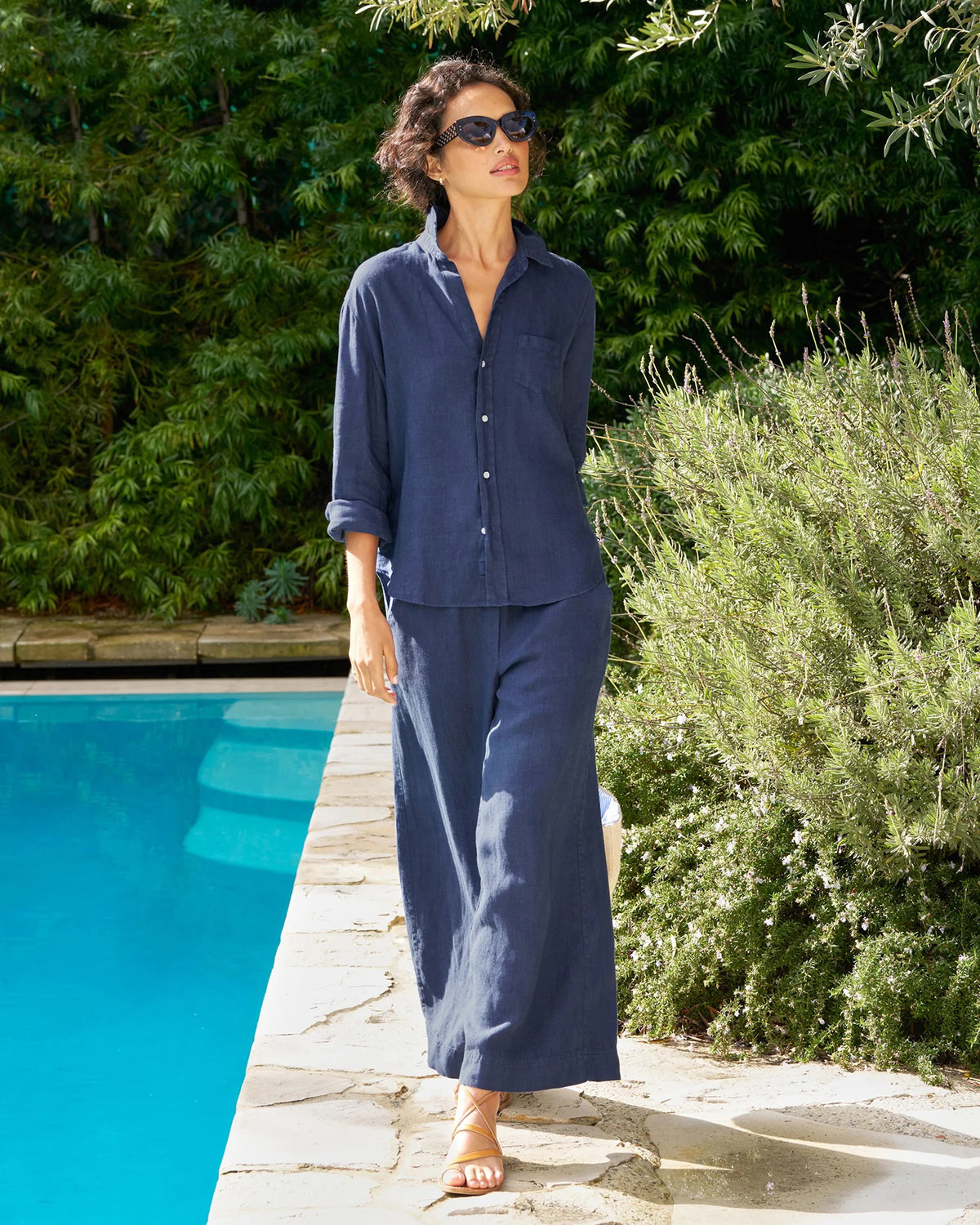 A woman in the FRANK &amp; EILEEN Eileen Relaxed Button Up Shirt in Linen from the Tuscany Getaway Set stands poolside among greenery, wearing wide-leg pants, sandals, sunglasses, and sporting short curly hair as she gazes upward.