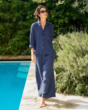 A woman in the FRANK & EILEEN Eileen Relaxed Button Up Shirt in Linen from the Tuscany Getaway Set stands poolside among greenery, wearing wide-leg pants, sandals, sunglasses, and sporting short curly hair as she gazes upward.