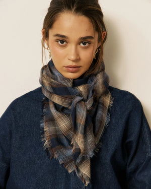 A woman stands indoors near a large window with natural light, wearing a dark denim jacket and pants, accessorized with the HARTFORD checked wool scarf in navy by HARTFORD.