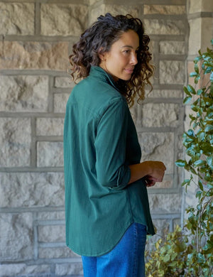 A woman with curly hair, smiling softly by a window indoors with one hand in her pocket, wears blue jeans and the FRANK & EILEEN Waverley Button Up Tee in Vintage Jersey Pine.
