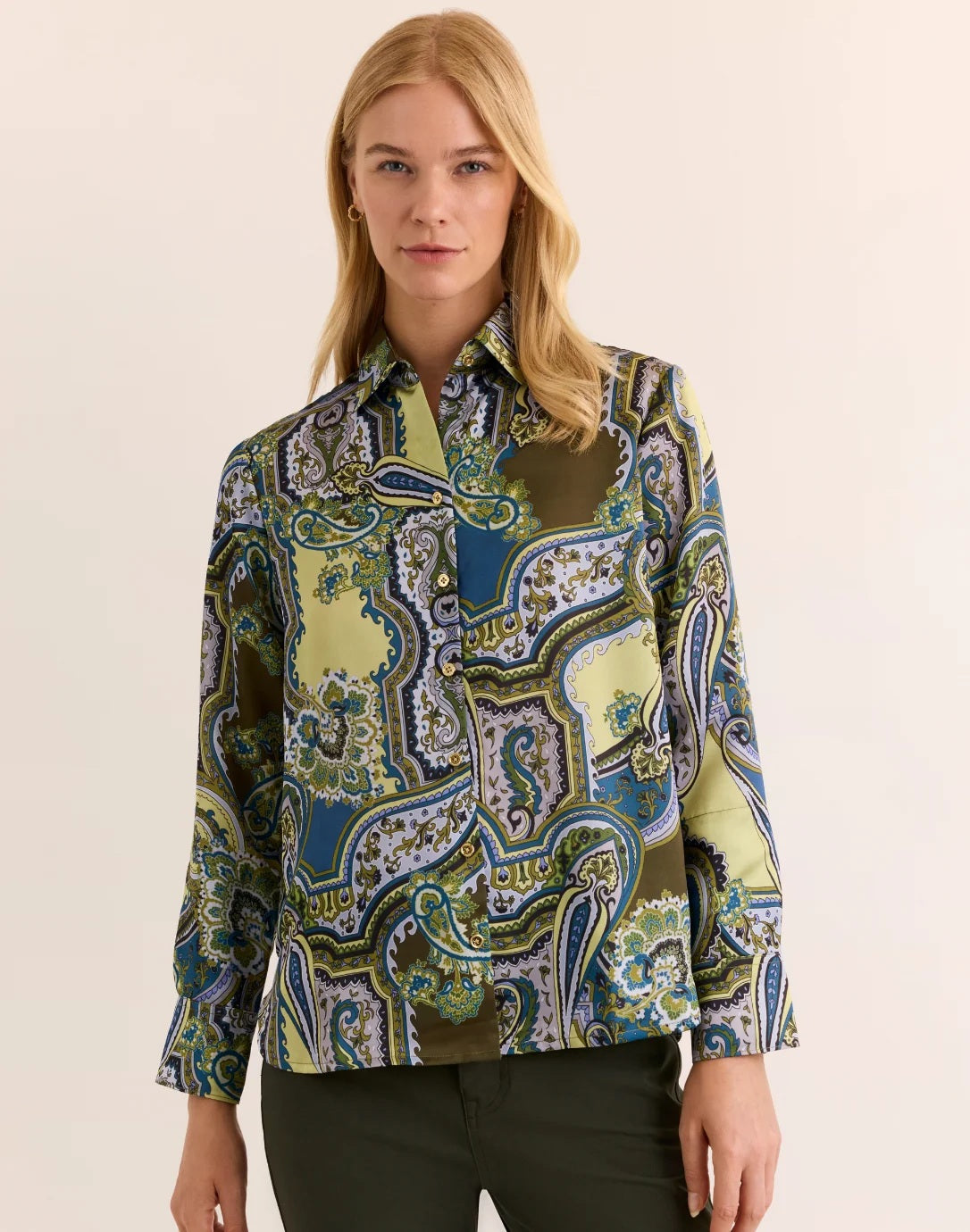 A woman with blonde hair wears the HINSON WU Margot Long Sleeve Paisley Shirt, a colorful button-up featuring green, blue, and yellow paisley patterns and crafted from luxurious Italian polyester for an elegant drape and silky feel. She pairs it with dark green pants and stands against a neutral background.