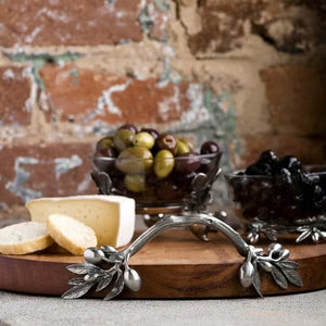 The OLIVE GROVE CHEESE BOARD by VAGABOND HOUSE is a round oak wood serving tray with a smooth surface and two ornately designed pewter-embellished handles on opposite sides. The handles feature intricate leaf and berry patterns, adding an elegant touch to the rustic tray.