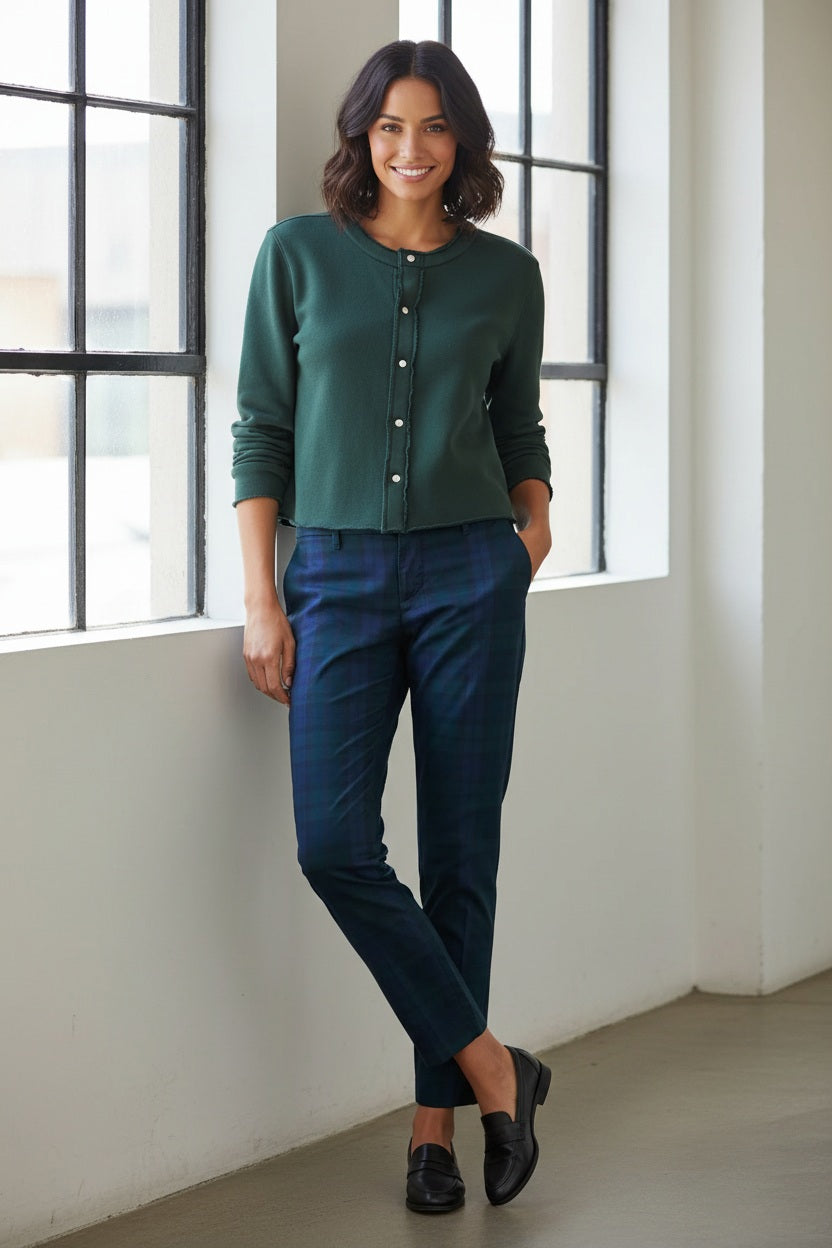 A woman with wavy dark hair stands by large windows, smiling as she wears the FRANK & EILEEN Jackie Sweatshirt Cardigan in Triple Fleece Pine from FRANK & EILEEN, paired with blue plaid pants and black loafers. One hand is tucked into her pocket and her legs are crossed.