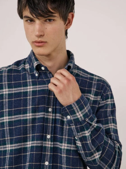 A young person with dark hair, wearing the HARTFORD - PITT CHECKED FLANNEL SHIRT IN BLUE AND GREEN by HARTFORD, looks at the camera while adjusting the shirt collar with one hand against a plain light background.