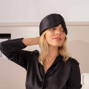 An older person with long gray hair wears a luxurious beige DROWSY - SATIN SLEEP MASK by DROWSY SLEEP CO, made of mulberry silk. In a matching satin top, they stand against a plain white background, ready for beauty sleep.