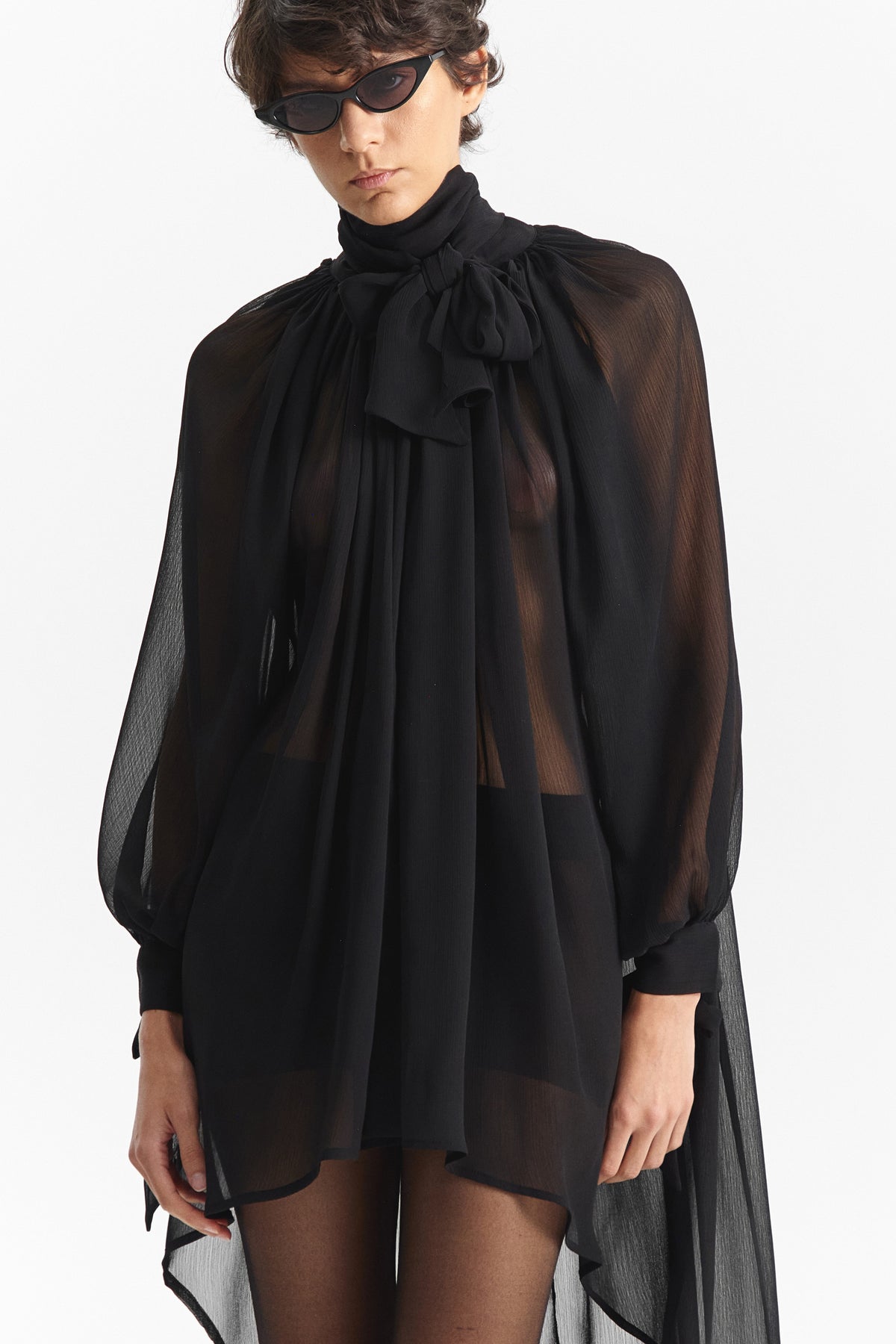 The ELEH - THE CHIFFON TIE NECK TOP by ELEH is a sheer black chiffon blouse with long sleeves, a high neckline, and an oversized bow tie at the collar—an elegant evening statement inspired by classic blouses.