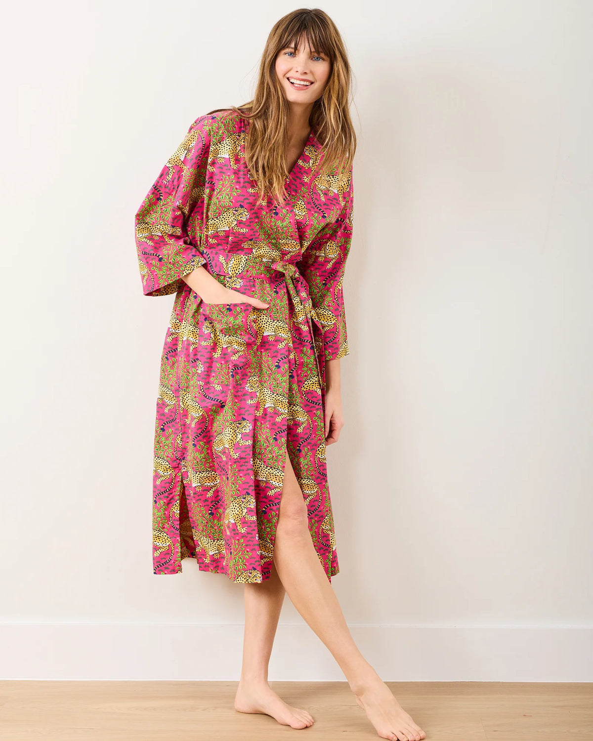 A person smiles while posing in the PRINTFRESH - BAGHEERA COTTON ROBE, a luxurious floral-patterned piece made from organic cotton with bold pink, yellow, and green designs. They stand barefoot against a plain, neutral background.