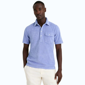 A person in a periwinkle Criquet Terrycloth Players Shirt and white shorts stands facing away from the camera against a plain beige background.