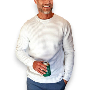 A smiling man with a short beard wears the CRIQUET - QUILTED CREW SWEATSHIRT IN OFF-WHITE by CRIQUET, holding a green can indoors near wood-paneled walls, a window, and a wooden chair.