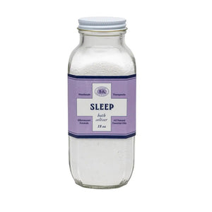 A clear glass jar labeled "BAUDELAIRE - SLEEP BATH SELTZER" features a purple label with ingredients, a barcode, and essential oils. Filled with granular white bath salts, its decorative border suggests instructions for a calming soak.
