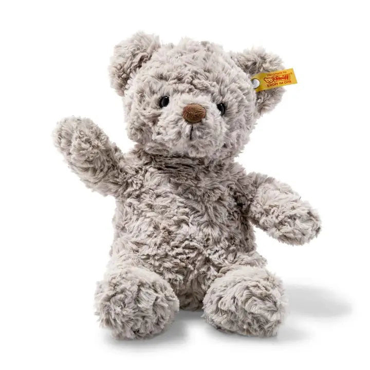 A fluffy, light gray STEIFF - HONEY TEDDY BEAR with a brown nose and black eyes sits upright. Its left arm is raised in a teaching grip, and a small yellow tag with red lettering is attached to its left ear. The bear's fur appears soft and shaggy.