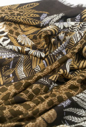 A person with shoulder-length gray hair stands facing the camera, wearing a white shirt and blue jeans. Adorning their neck is an INOUI EDITIONS - ROUSSEAU WOOL SCARF by INOUI, featuring an artisan-made brown fabric with a floral and animal pattern. The background is plain white.