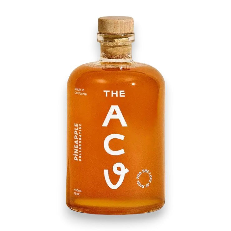 A clear, round glass bottle with a cork stopper, filled with golden liquid. The label reads &quot;APPLE CIDER VINEGAR&quot; in large white letters. Smaller text indicates &quot;PINEAPPLE COLLABORATIVE&quot; and &quot;Made in California&quot;. The bottle has a minimalist design and contains 450ml of probiotic-rich apple cider vinegar made from organic apples.