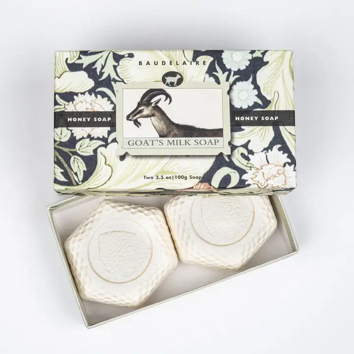 A BAUDELAIRE GOATS MILK HONEY SOAP 3.5OZ 2 BAR GIFT BOX features a floral, goat-illustrated box labeled "Honey Soap." Inside are two hexagonal, white moisturizing bars with embossed designs—ideal for sensitive skin.