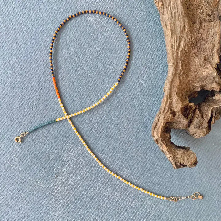 The ALICE RISE - MOSAIC NECKLACE IN GOLDEN ERA by ALICE RISE is a handmade beaded choker in yellow, orange, brown, and green tones, shown arranged in a loop on a textured blue surface beside driftwood.