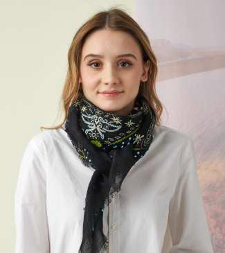 A young woman with light brown hair wears the STORIATIPIC Nelly Square Cashmere and Wool Embroidered Bandana Scarf, paired with a light gray top, standing before a softly colored nature-themed backdrop.