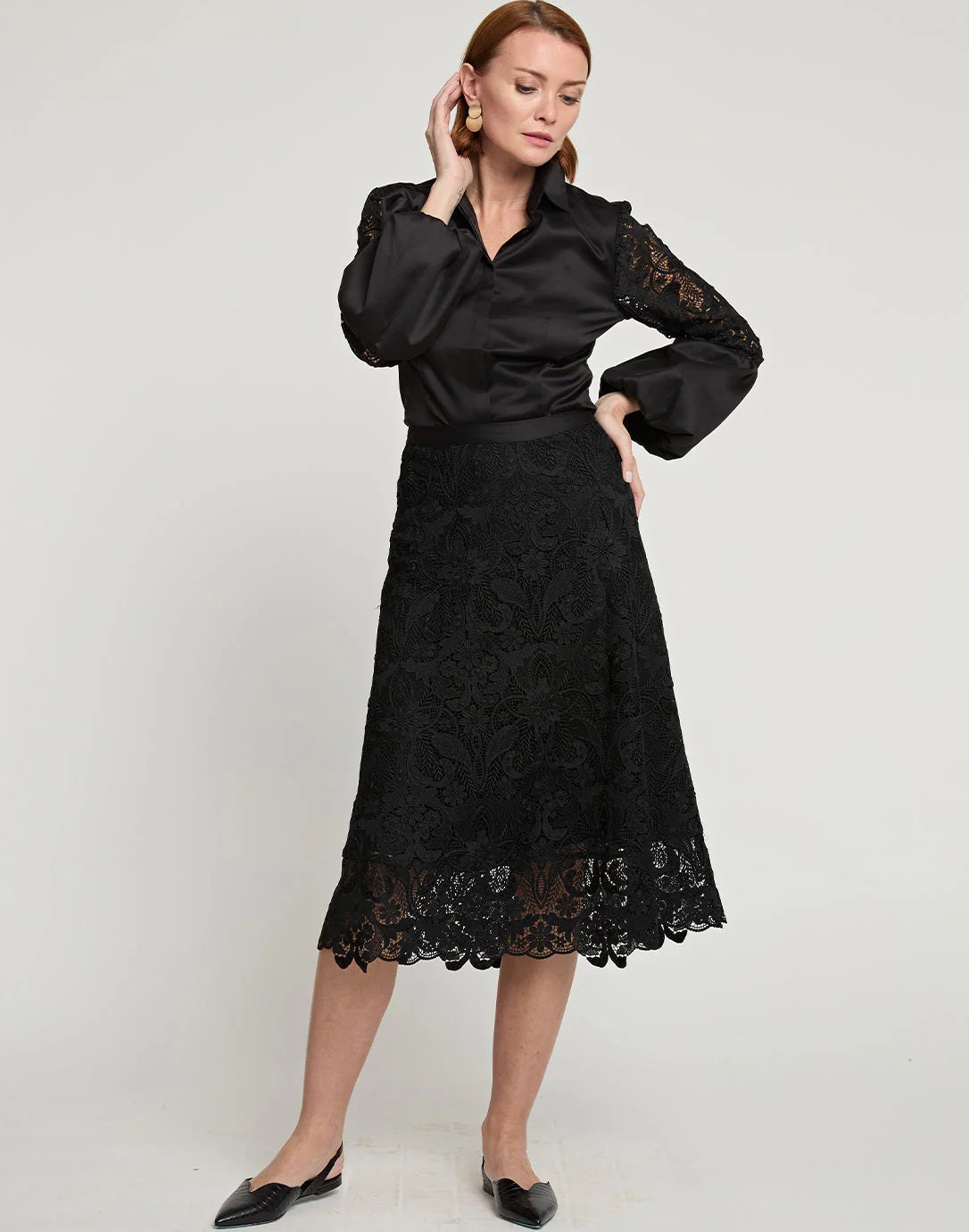 A woman stands against a plain backdrop wearing a black blouse featuring lace sleeves paired with the GLORIA LACE SKIRT from HINSON WU. Her red hair provides an elegant contrast to her attire as she touches her earring with one hand, completing the ensemble with stylish black shoes.