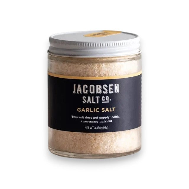A glass jar of Jacobsen Salt Co. Garlic Salt Infused Sea Salt, featuring a metal lid and a black-and-gold label, filled with coarse garlic-infused sea salt made from premium kosher salt.