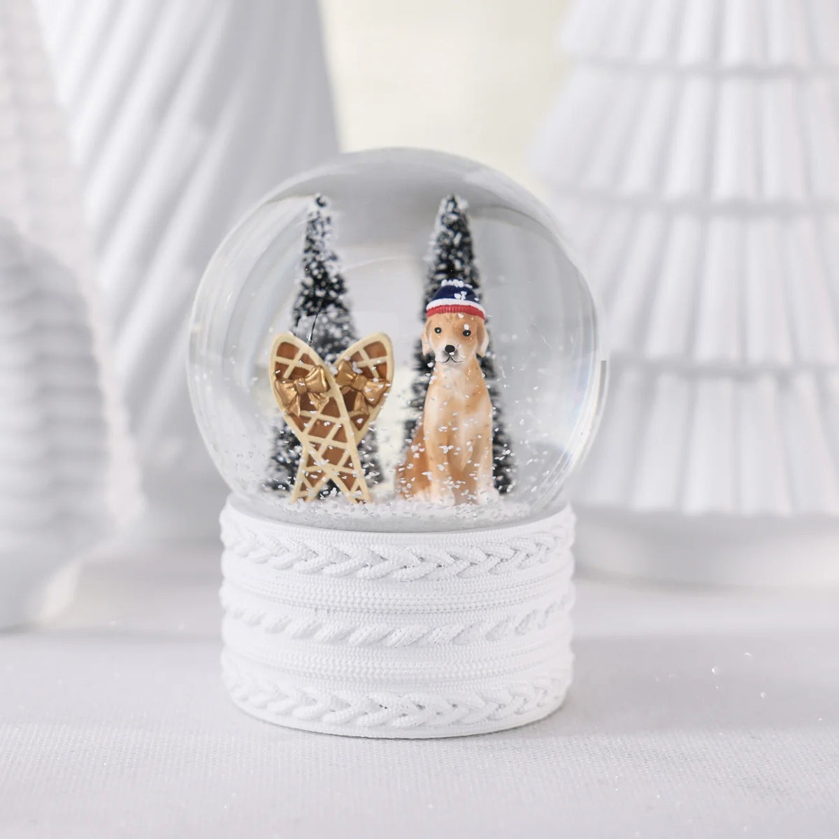 A ZODAX SNOW SHOEING MOUNTAIN DOG SNOW GLOBE featuring a resin glass globe with a dog in a hat, snowshoes, and two pine trees, set on a white cable-knit textured base.