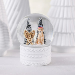 A ZODAX SNOW SHOEING MOUNTAIN DOG SNOW GLOBE featuring a resin glass globe with a dog in a hat, snowshoes, and two pine trees, set on a white cable-knit textured base.