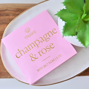 A pink box labeled “MUSEE - CHAMPAGNE & ROSE MINI BATH SALT SOAK” in gold lettering, with a small pile of white bath salt crystals displayed in front.