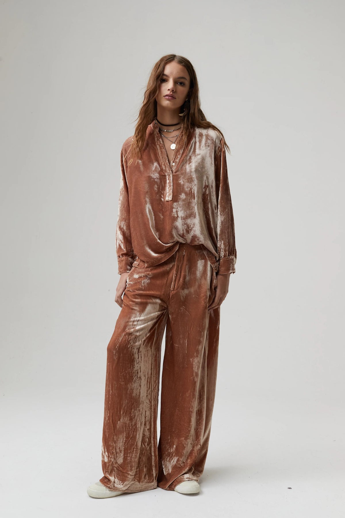 A person with long wavy hair stands against a plain background wearing a mustard yellow, pure silk velvet long-sleeve top and matching, high-waisted LANHTROPY - THE LUNA VELVET PANT by LANHTROPY. They are accessorized with layered necklaces, a brown belt, and flat sandals.