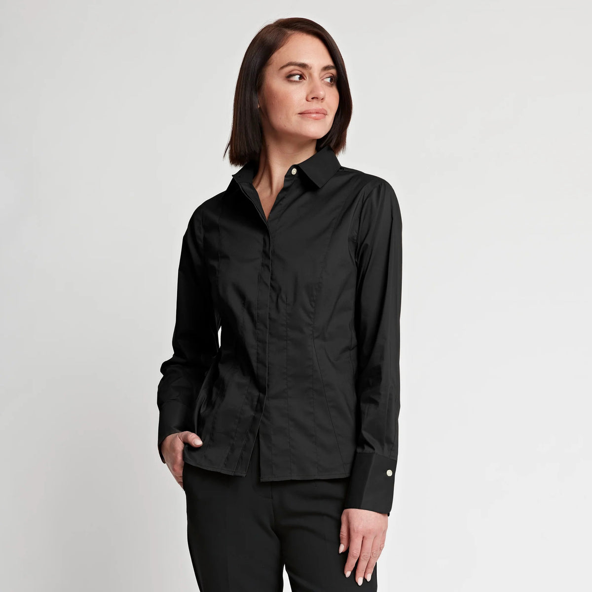 A woman with straight brown hair, wearing the HINSON WU - FRANCIS FITTED SHIRT—a tailored black button-up crafted from stretch polished cotton by HINSON WU—and black pants, stands with one hand in her pocket, gazing off to the side against a plain white background.