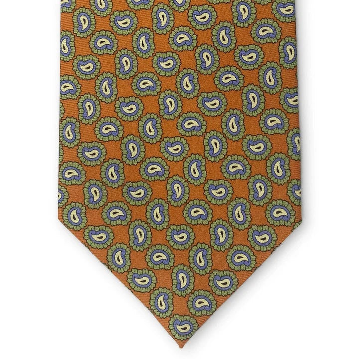 Introducing the PAGE SILK TIE IN ORANGE by BIRD DOG BAY. This expertly handmade New York tie features a vibrant orange base adorned with a repeating blue and yellow paisley pattern edged in green. With its pointed tip and solid white background, the PETER-BLAIR tie elevates classic design for a modern, sophisticated look.