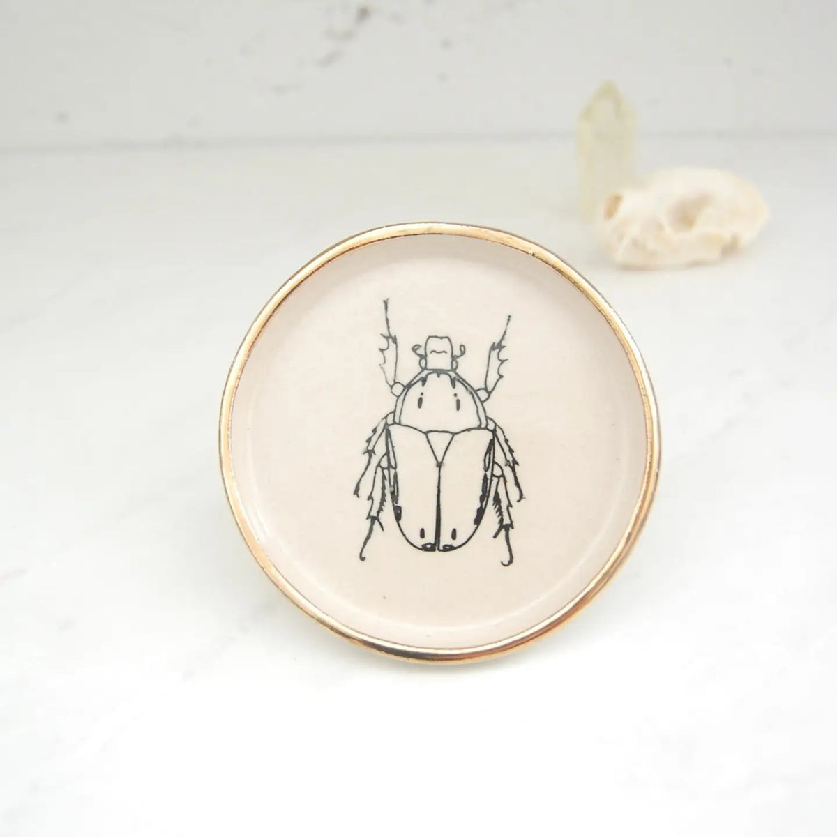 The COMPOST DESIGN - BEETLE DISH 4&quot; by COMPOST DESIGN showcases a black beetle illustration at its center, surrounded by a 22k gold rim. Set on white, it features an abstract shell design background—perfect for decoration or as part of a treasured collection.