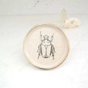 The COMPOST DESIGN - BEETLE DISH 4" by COMPOST DESIGN showcases a black beetle illustration at its center, surrounded by a 22k gold rim. Set on white, it features an abstract shell design background—perfect for decoration or as part of a treasured collection.