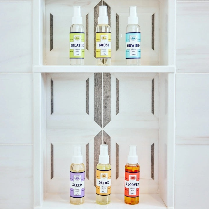 Six small bottles labeled Breathe, Boost, Unwind, Sleep, Detox, and Recover—each filled with essential oil shower mists like uplifting lemon and ginger—are arranged on white shelves in a tiled shower niche with a geometric gray and white pattern. Create your own aromatherapy steam room right at home.