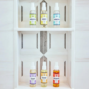 Six small bottles labeled Breathe, Boost, Unwind, Sleep, Detox, and Recover—each filled with essential oil shower mists like uplifting lemon and ginger—are arranged on white shelves in a tiled shower niche with a geometric gray and white pattern. Create your own aromatherapy steam room right at home.