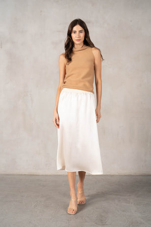 A woman in a sleeveless black dress with a wide belt stands against a plain backdrop, holding a large black handbag. She wears black shoes and layers the ROSE AND CROWN - SATIN RIBBED SKIRT by ROSE & CROWN under her dress.