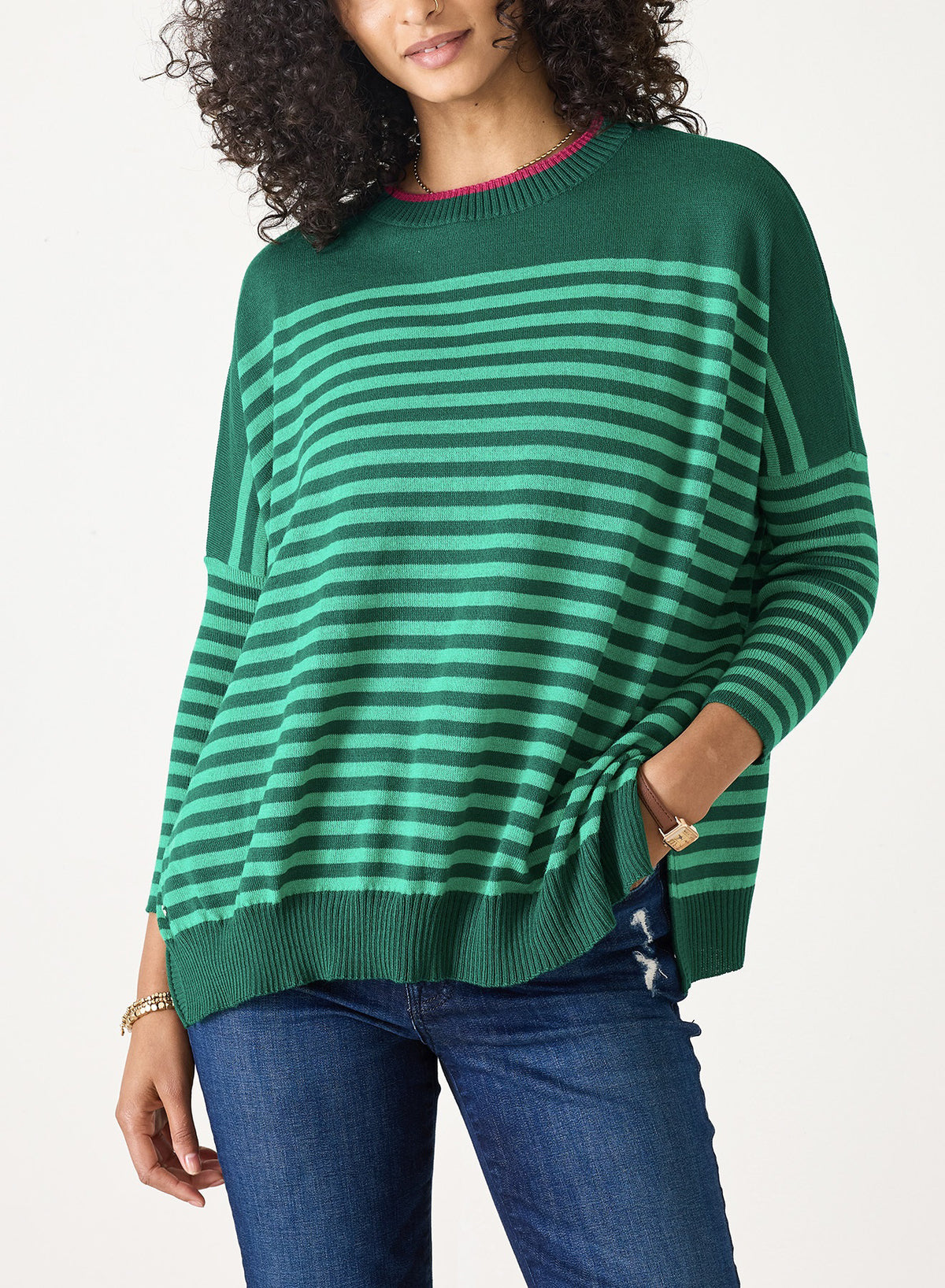 A person with curly hair wears the MERSEA Catalina Modern Crewneck Sweater in Spruce/Emerald and blue jeans, standing with one hand in their pocket against a plain white background.