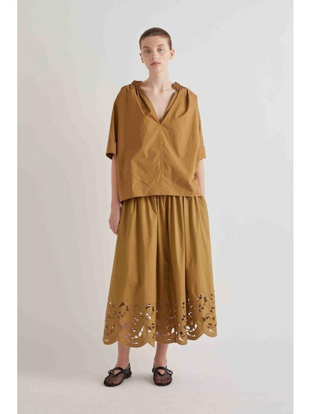 A woman stands against a plain background wearing the MEIMEIJ Gathered Neck Blouse in oak with short sleeves, paired with a matching midi skirt featuring a cut-out floral hem and black sandals.