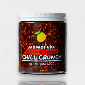 A jar of MOMOFUKU - EXTRA SPICY CHILI CRUNCH is shown against a white background. The jar contains 150 grams (5.3 ounces) of smoky spiciness, with a vibrant red color and visible Mexican chilis, highlighted by a yellow peach graphic above the brand name, MOMOFUKU.