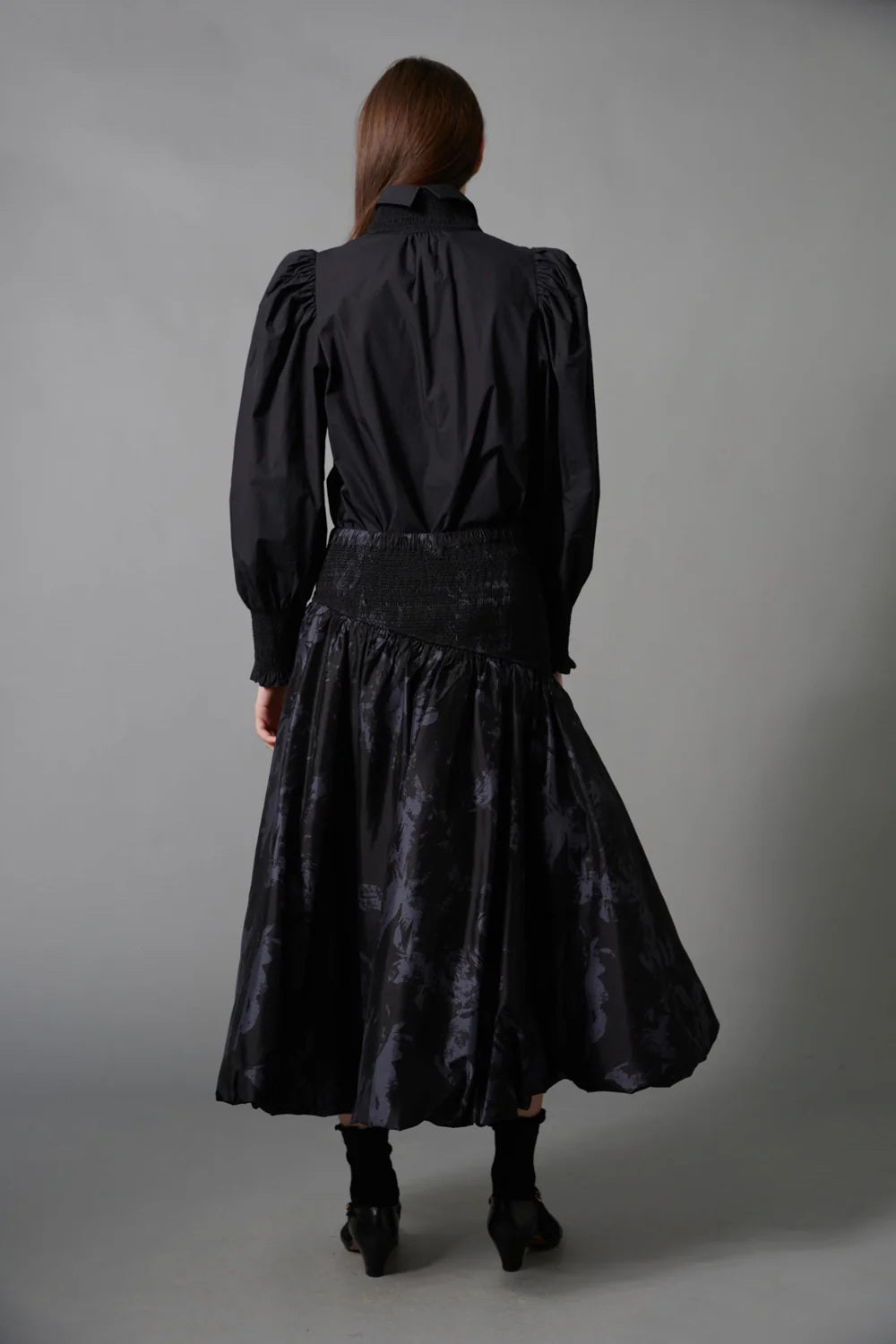 A woman with long brown hair faces away, wearing the HOPE FOR FLOWERS Smocked Shirt in Black with puffed sleeves and paired with a dark floral skirt, standing before a gray backdrop.