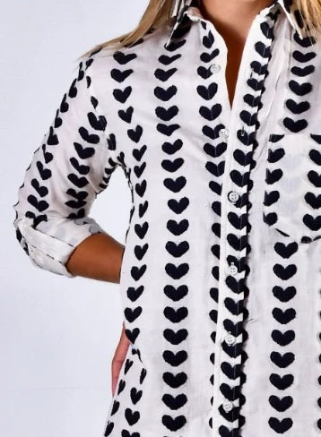 A woman stands in a sunlit room wearing the EMILY PHILLIPS - BLACK HEARTS GIRLFRIEND BUTTON DOWN SHIRT from EMILY PHILLIPS—its playful heart pattern and cotton fabric paired with blue jeans and brown boots. Sunlight pours through large windows, casting shadows across the floor.